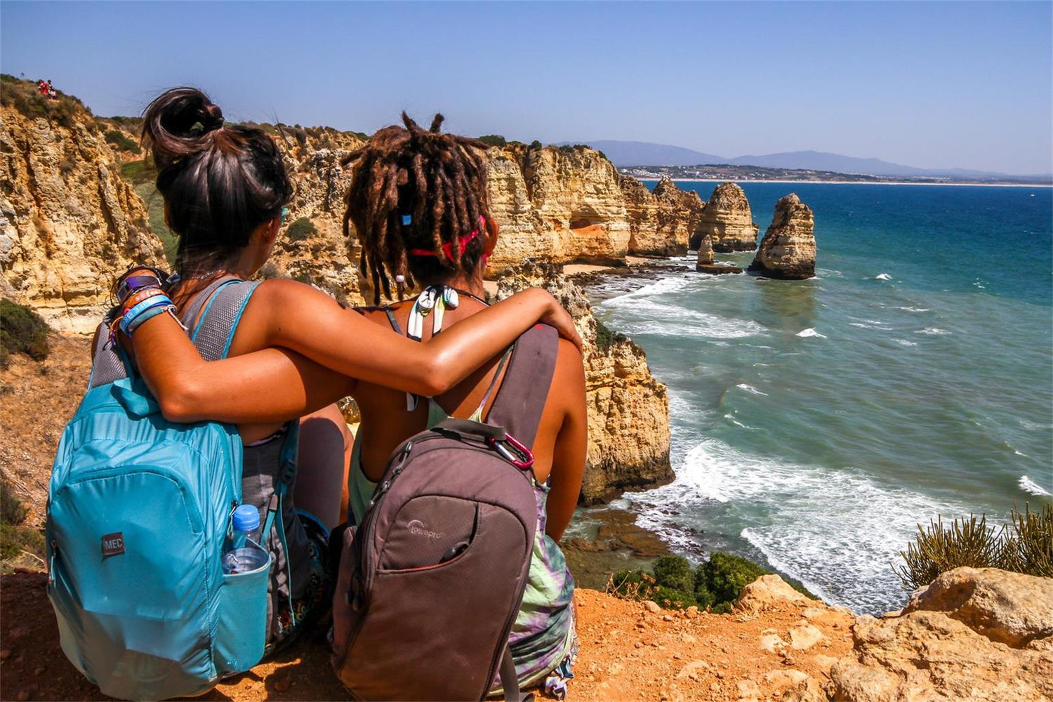 Two women with backpacks sitting on cliff edge, gazing at ocean. Join Apexel Partners Affiliate Program!